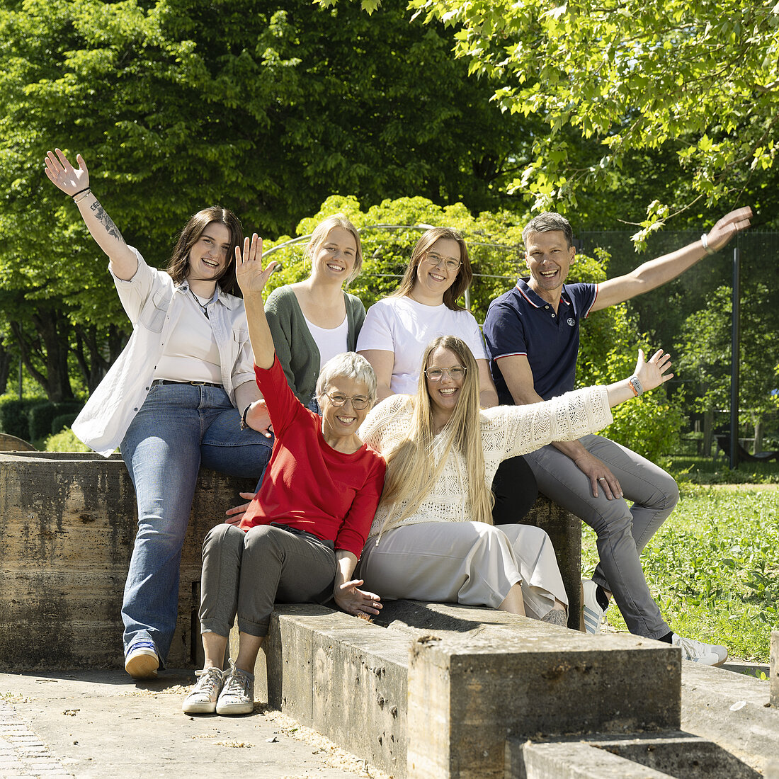 Sechs Menschen unterschiedlicher Altersgruppen sitzen auf Steinbänken im Park, einige mit erhobenen Armen.